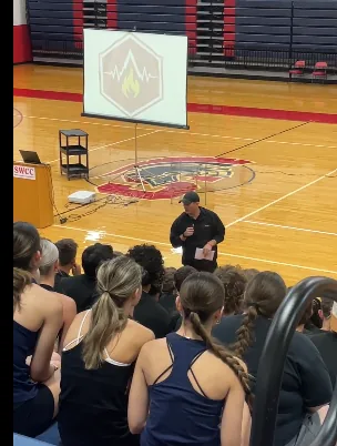 Trevor Conner speaking with athletes in a gym, with the Hearts and Minds symbol projected behind him